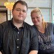 A man and a woman sitting together in a casual setting, with the woman smiling and the man looking directly at the camera. A man and a woman sitting together in a casual setting, with the woman smiling and the man looking directly at the camera.