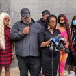 A group of people stands together at a press conference, with one woman speaking into a microphone while holding a piece of paper, as others listen in support. A group of people stands together at a press conference, with one woman speaking into a microphone while holding a piece of paper, as others listen in support.