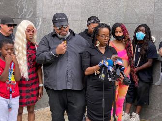 A group of people stands together at a press conference, with one woman speaking into a microphone while holding a piece of paper, as others listen in support.