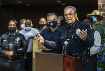 A police official speaks at a podium during a public event, with officers and community members present in the background.