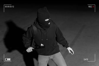 A person wearing a ski mask and dark clothing holding a weapon in a dimly lit environment, suggesting a robbery scenario.