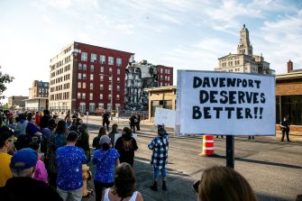 Crowd gathered near the site of an apartment building collapse in Davenport, Iowa, holding a sign that reads "Davenport deserves better."