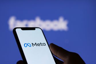 A hand holding a smartphone displaying the Meta logo, with the Facebook logo blurred in the background.