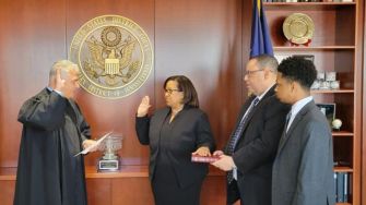 A federal judge administering an oath of office to a newly appointed judge, surrounded by family and holding a book, with an official seal in the background.