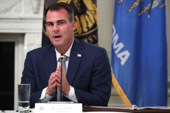 Governor Kevin Stitt of Oklahoma speaking during a meeting, with state flags in the background.