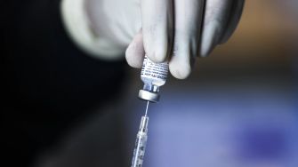 A gloved hand holding a vial of COVID-19 vaccine while preparing a syringe for vaccination.