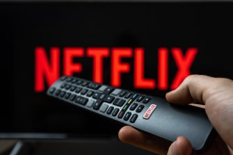 A hand holding a remote control in front of a screen displaying the Netflix logo.