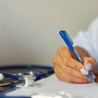 A close-up of a person's hand writing on paper with a pen, alongside a stethoscope and a pot with a cactus in the background. A close-up of a person's hand writing on paper with a pen, alongside a stethoscope and a pot with a cactus in the background.