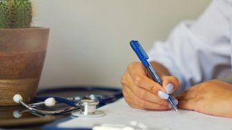 A close-up of a person's hand writing on paper with a pen, alongside a stethoscope and a pot with a cactus in the background.