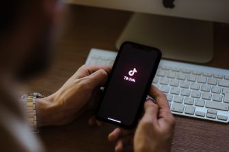 A person holding a smartphone displaying the TikTok logo, with a computer keyboard in the background.