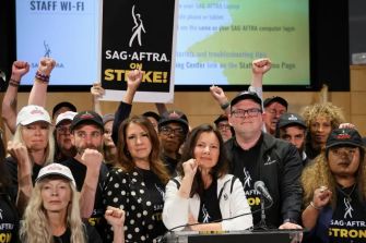 A group of actors and union representatives at a press conference, all showing solidarity with raised fists and holding signs that read "SAG-AFTRA ON STRIKE!"