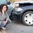 A woman is on the phone looking concerned after a car accident, with a damaged vehicle in the foreground. A woman is on the phone looking concerned after a car accident, with a damaged vehicle in the foreground.