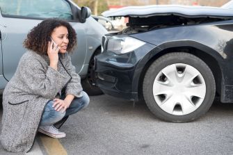 A woman is on the phone looking concerned after a car accident, with a damaged vehicle in the foreground.