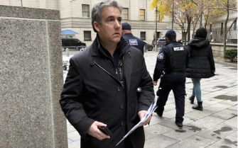 Michael Cohen walking outside a courthouse, holding documents, with security personnel in the background.