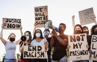 A diverse group of protesters holding signs with messages about justice, racism, and political dissent, while some use megaphones to amplify their voices.