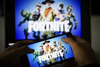 A person holding a smartphone showing the game "Fortnite," with the game's logo and characters displayed on the screen.