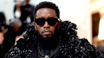 Sean "Diddy" Combs wearing sunglasses and a detailed black outfit adorned with roses, looking serious amidst a blurred background.
