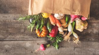 A paper bag spilling over with fresh vegetables and herbs on a wooden surface.