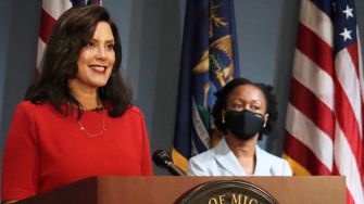 Governor Gretchen Whitmer speaking at a press conference with another official, with flags displayed in the background.