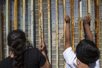 Two children look through gaps in a border wall, highlighting the difficult crossing experience for migrants.