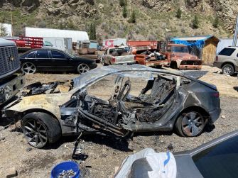 A severely damaged Tesla Model 3, burnt and wrecked, in a salvage yard after a fatal crash.