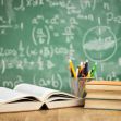 A classroom setting featuring an open book, stacked books, and a cup of colorful pencils in front of a green chalkboard with mathematical equations. A classroom setting featuring an open book, stacked books, and a cup of colorful pencils in front of a green chalkboard with mathematical equations.