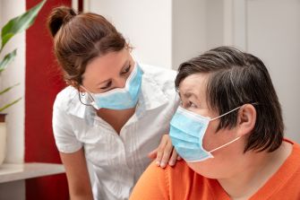 A caregiver wearing a mask comforts a person with a disability, both indoors.