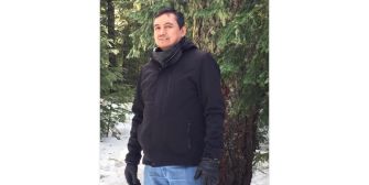 A man standing outdoors in a snowy area, wearing a black jacket and gloves, with trees in the background.