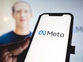 A hand holding a smartphone displaying the Meta logo on the screen, with a blurred image of a person in the background.