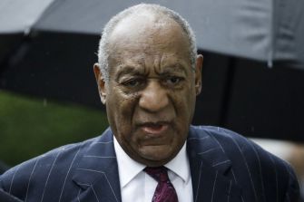 Bill Cosby walking under an umbrella, appearing serious during a legal proceeding.