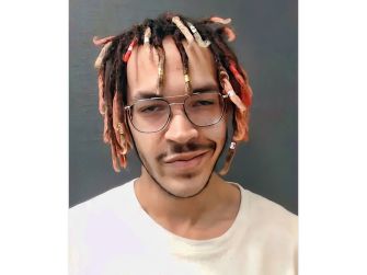 A young man with colorful dreadlocks and glasses poses for a mugshot against a dark background.