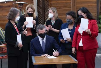California Governor Gavin Newsom signs a new COVID-19 sick leave law, surrounded by officials and supporters, all wearing masks.