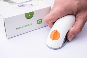 A person holding a Dexcom G6 Continuous Glucose Monitoring device with its packaging in the background.