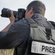A freelance journalist wearing a vest labeled "PRESS" is photographing an outdoor scene. A freelance journalist wearing a vest labeled "PRESS" is photographing an outdoor scene.