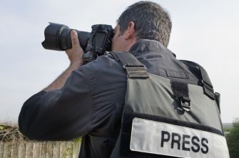A freelance journalist wearing a vest labeled "PRESS" is photographing an outdoor scene.