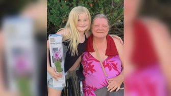 A young girl standing next to an older woman in a wheelchair, both smiling, outdoors with green foliage in the background.