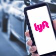 A person holding a smartphone displaying the Lyft app, with blurred cars in the background. A person holding a smartphone displaying the Lyft app, with blurred cars in the background.
