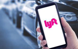 A person holding a smartphone displaying the Lyft app, with blurred cars in the background.