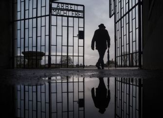 A silhouette of a person walking through an open gate with the words "ARBEIT MACHT FREI" (Work Sets You Free) reflected in a puddle on the ground.