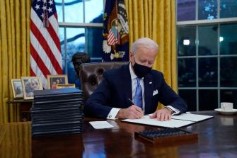 President Joe Biden signing executive orders in the Oval Office on his first day in office, wearing a mask and surrounded by stacks of documents.
