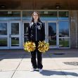 A teenage girl stands outside a school building holding gold pom-poms while wearing a cheerleading outfit, symbolizing her involvement in cheerleading. A teenage girl stands outside a school building holding gold pom-poms while wearing a cheerleading outfit, symbolizing her involvement in cheerleading.
