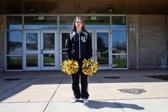 A teenage girl stands outside a school building holding gold pom-poms while wearing a cheerleading outfit, symbolizing her involvement in cheerleading.