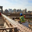A worker in safety gear performs maintenance on the Brooklyn Bridge with the city skyline in the background. A worker in safety gear performs maintenance on the Brooklyn Bridge with the city skyline in the background.