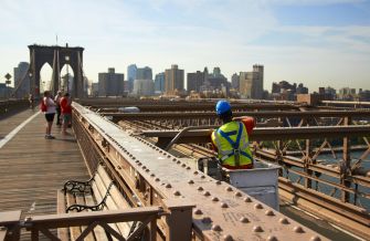 A worker in safety gear performs maintenance on the Brooklyn Bridge with the city skyline in the background.