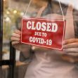 A person holding a red "CLOSED DUE TO COVID-19" sign in front of a shop window. A person holding a red "CLOSED DUE TO COVID-19" sign in front of a shop window.