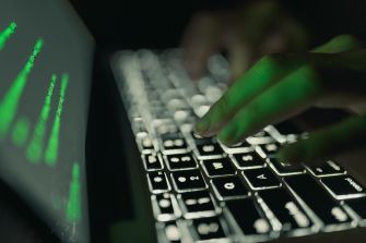 A close-up of hands typing on a laptop keyboard with green code displayed on the screen, illustrating themes of cybersecurity and data breaches.