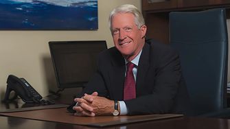 A businessman seated at a desk, smiling and wearing a suit, with a phone and computer in the background.