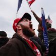 A man with a beard wearing a red cap and a brown jacket is shouting among a crowd of people holding flags. A man with a beard wearing a red cap and a brown jacket is shouting among a crowd of people holding flags.