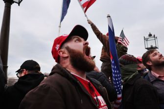 A man with a beard wearing a red cap and a brown jacket is shouting among a crowd of people holding flags.