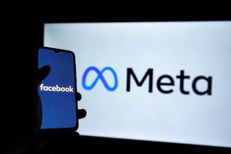 A person holding a smartphone displaying the Facebook logo in front of a screen with the Meta logo.